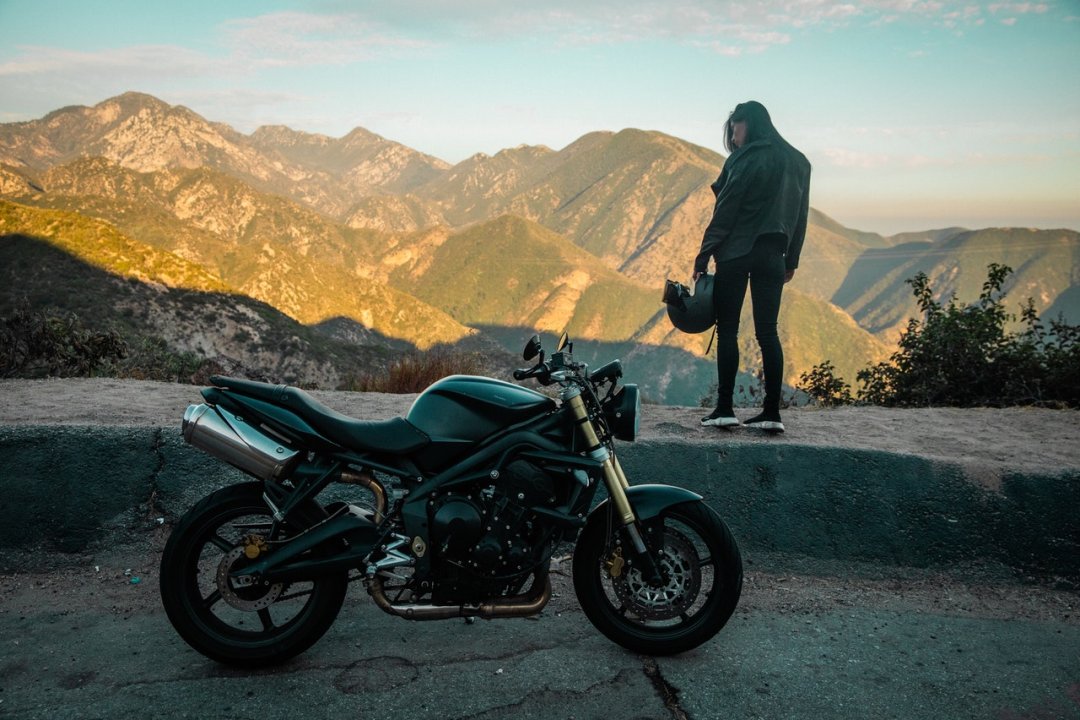Femme debout à côté d’une moto roadster de couleur noire garée au bord d’une route de montagne